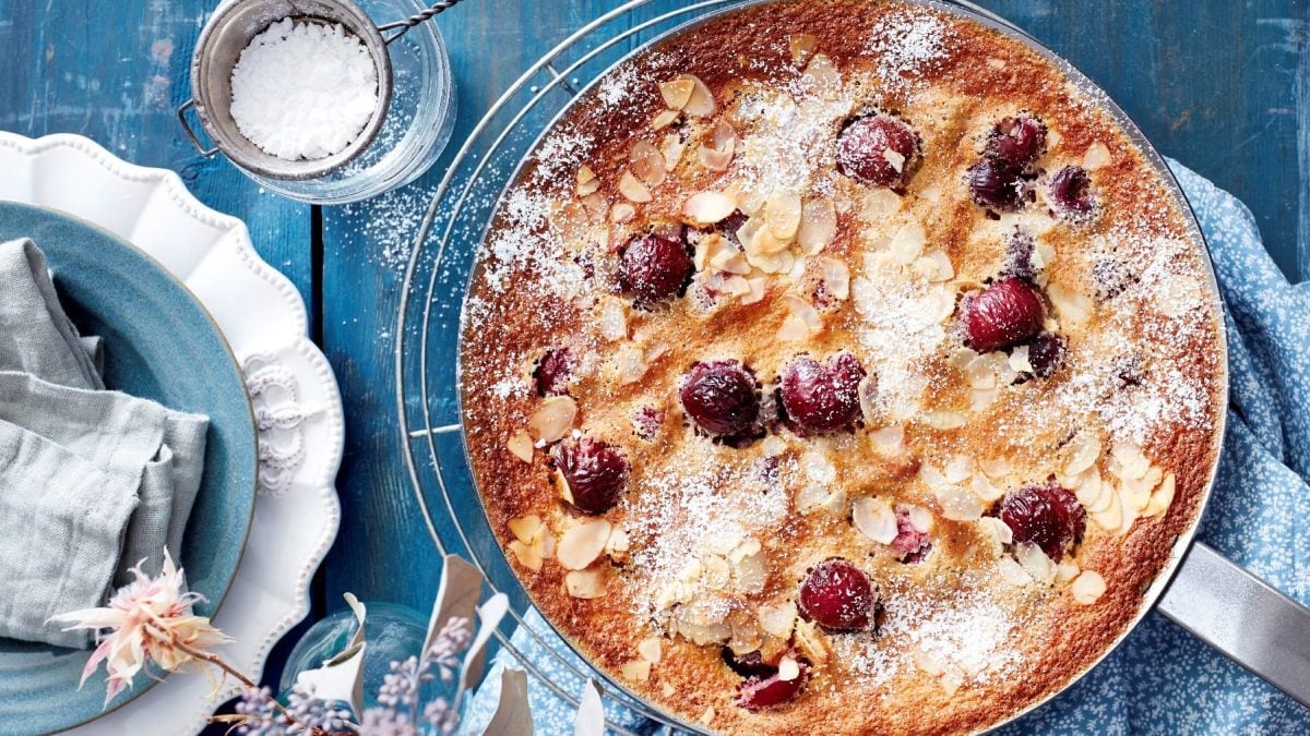 Kirsch-Clafoutis, bestehend aus einem Pfannkuchenteig und frischen Kirschen, gegossen in eine Kuchenform und ausgebacken, und bestreut mit Mandeln und Puderzucker, serviert auf einem blauen Tisch.