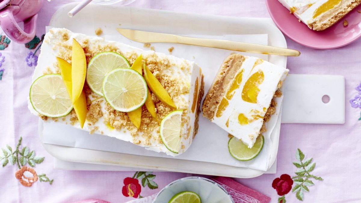 Auf einem Teller steht ein Mango-Kokos-Cheesecake. Er ist mit Limetten und Mango belegt und mit Streuseln bestreut.