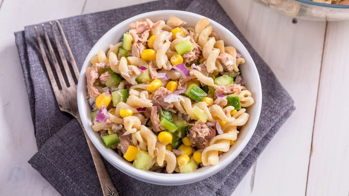 Nudelsalat mit Thunfisch, Gurke, Mais und Zwiebel in weißer Schale auf grauem Tuch.