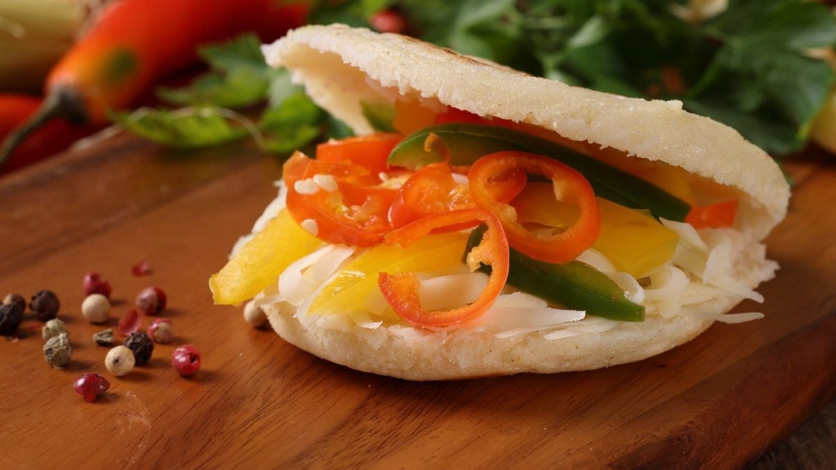 Arepas mit Chili-Kraut und unten Paprika-Streifen auf Holztisch. Daneben bunter Pfeffer, Chilis und Petersilie.
