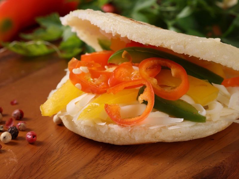 Arepas mit Chili-Kraut und unten Paprika-Streifen auf Holztisch. Daneben bunter Pfeffer, Chilis und Petersilie.
