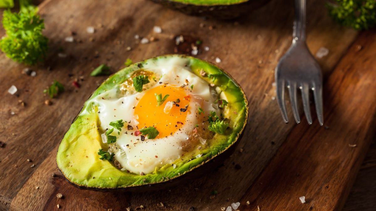 Eine Avocadohälfte mit einem Spiegelei gefüllt auf einem rustikalen Holzbrett. Daneben einiges an Kräutern und Gewürzen.