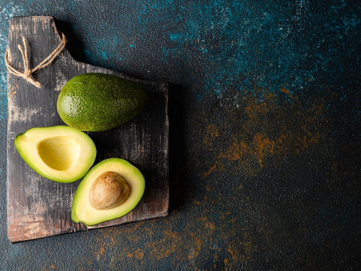 Ein dunkles Schneidebrett mit einer ganzen und einer aufgeschnittenen Avocado. Der Tisch ist rostfarben mit blauer Schraffur.