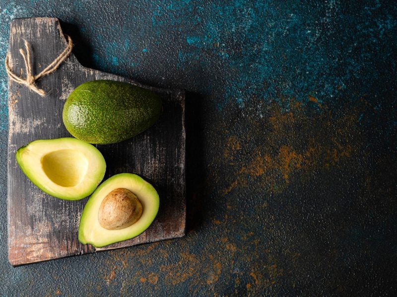 Ein dunkles Schneidebrett mit einer ganzen und einer aufgeschnittenen Avocado. Der Tisch ist rostfarben mit blauer Schraffur.