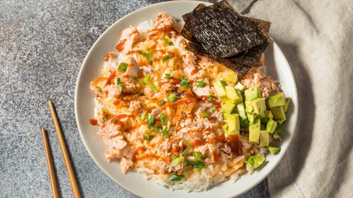 Ein Teller mit Bang Bang Salmon, Avocado und Nori-Blätter auf einem weißen Teller neben einem Paar Stäbchen.