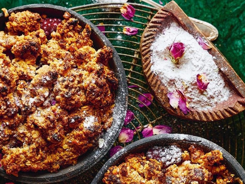 Ein birnen-brombeer-crumble steht auf einem Kuchengitter. Drum herum sind noch zwei Crumbles.