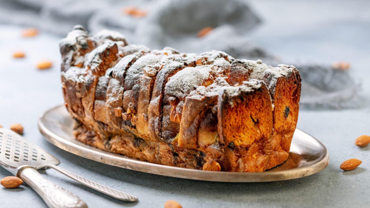 Brotpudding mit Ahornsirup, Banane und Nüssen auf weißem Teller. Im Bild Bestecke und Nüsse. Frontalansicht.
