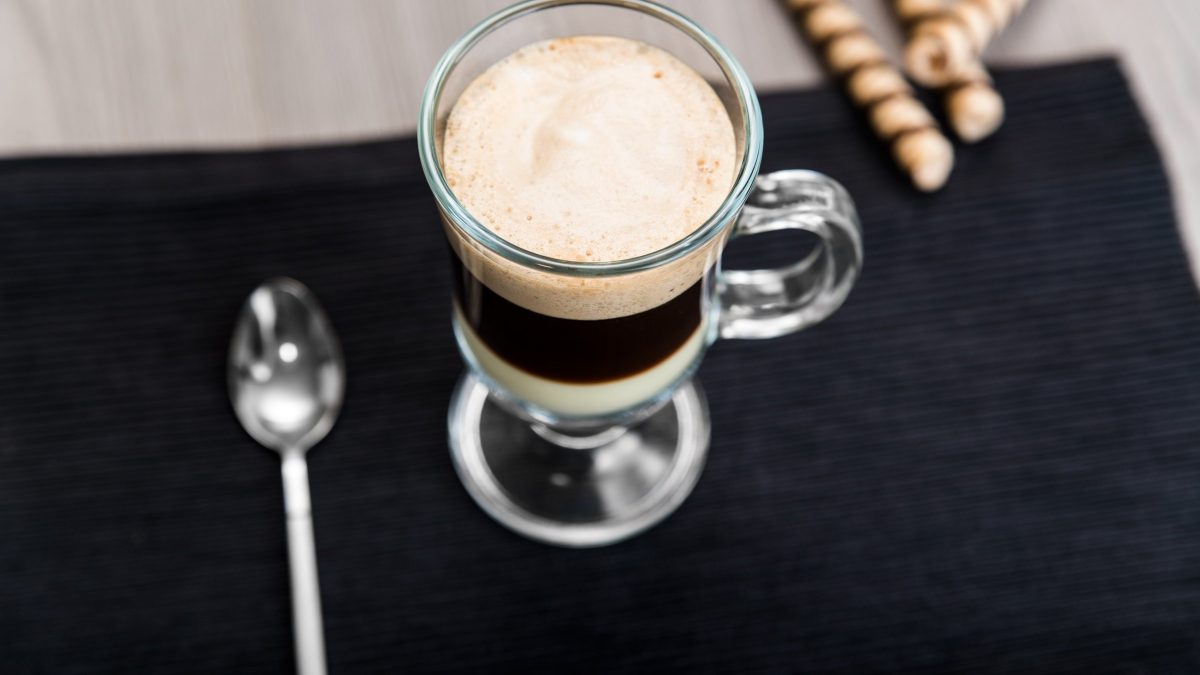 Café Bombón auf schwarzer Platte auf hellem Tisch. Daneben Metalllöffel und Kekse. Draufsicht.