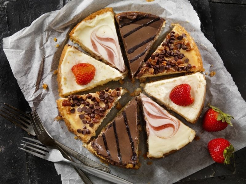 Verschiedene Cheesecake-Stücke liegen auf einem Stück Backpapier. Daneben liegen Erdbeeren und Gabeln.