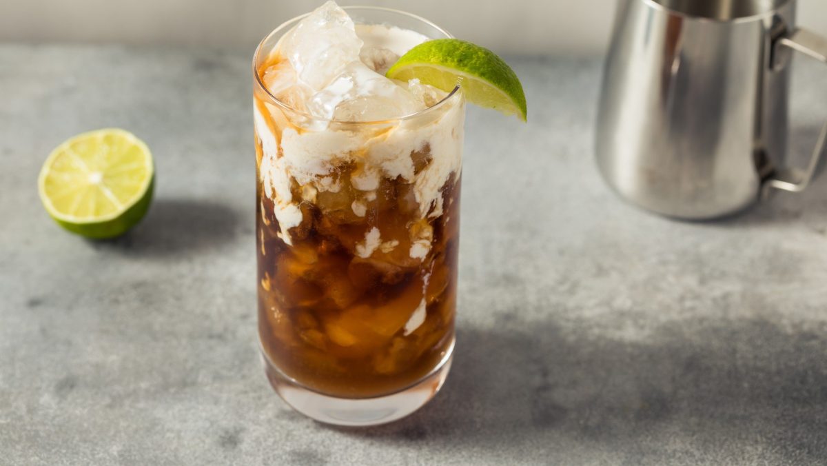 Dirty Soda in einem Glas auf grauem Untergrund. Daneben Limette und Sahnekännchen. Frontalansicht.