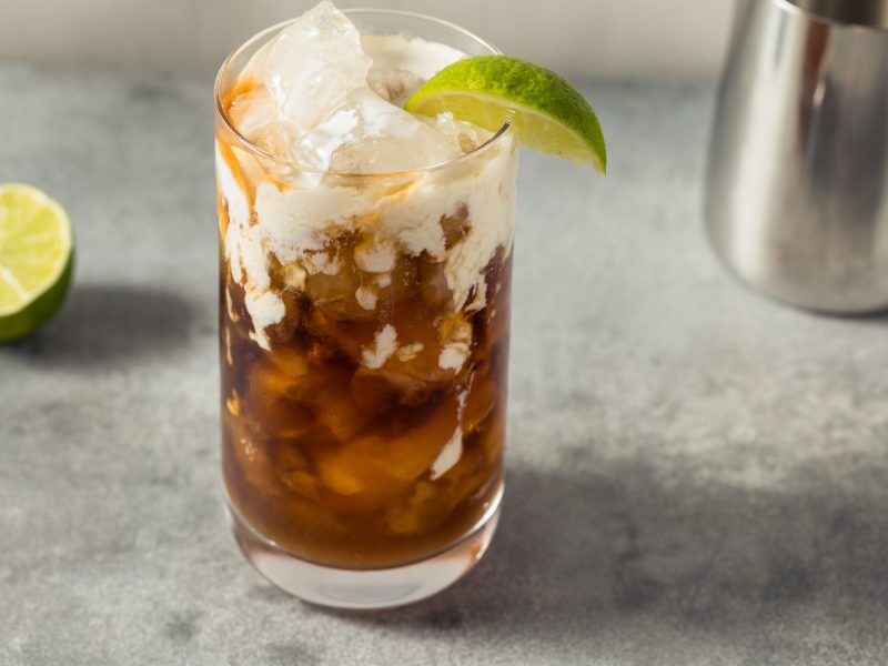Dirty Soda in einem Glas auf grauem Untergrund. Daneben Limette und Sahnekännchen. Frontalansicht.