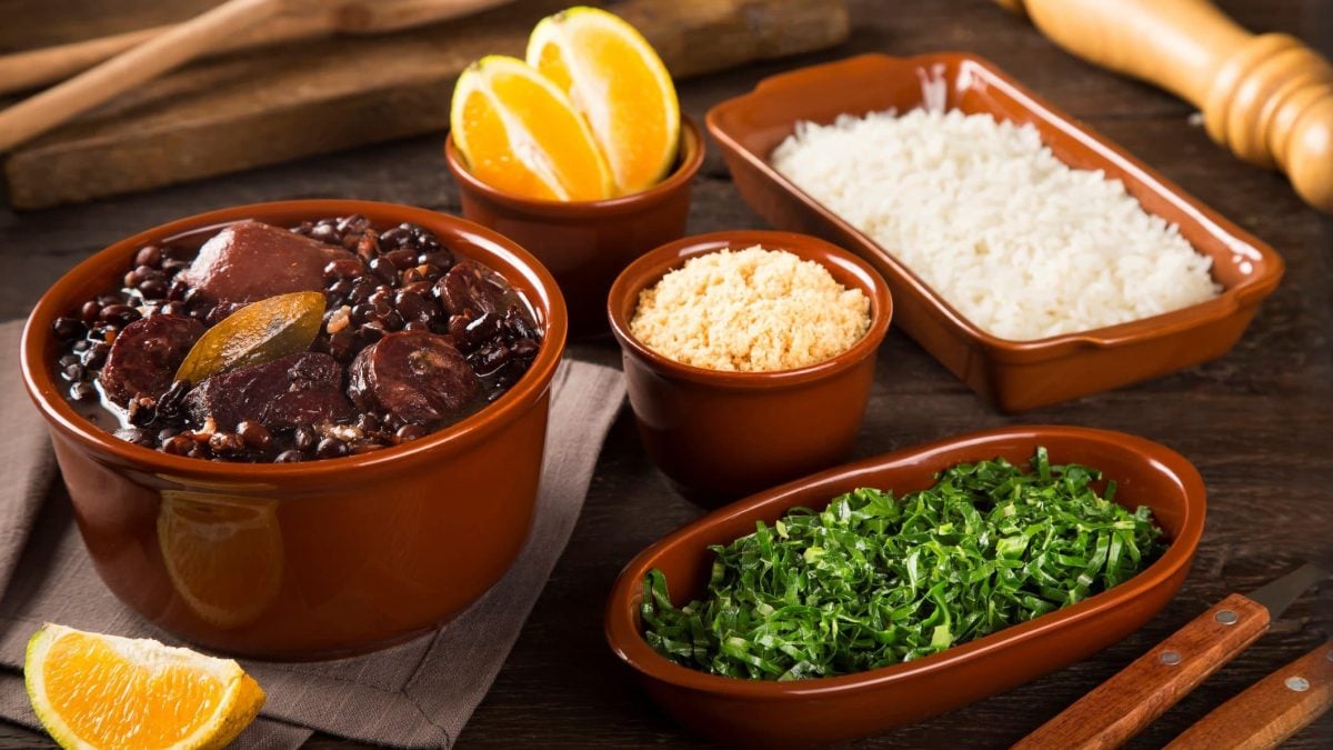 Feijoada in Tonform auf braunem Untergrund. Daneben Beilagen in Tonformen. Holz als Deko im Bild. Frontalansicht.