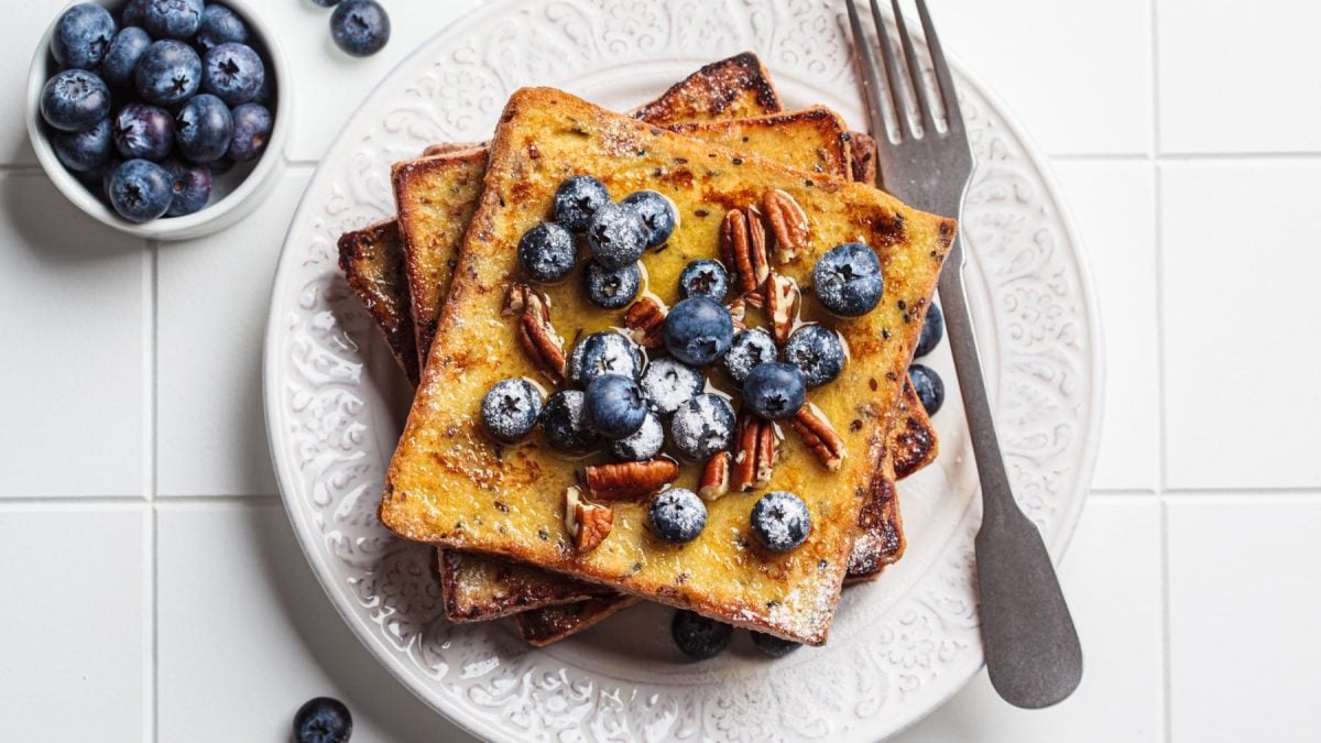 French Toast mit Heidelbeeren auf einem weißen Teller. Daneben liegt eine Gabel und eine Schale mit Heidelbeeren. Das ganze auf deinem gefliesten Untergrund.