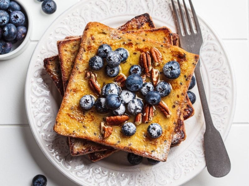 French Toast mit Heidelbeeren auf einem weißen Teller. Daneben liegt eine Gabel und eine Schale mit Heidelbeeren. Das ganze auf deinem gefliesten Untergrund.