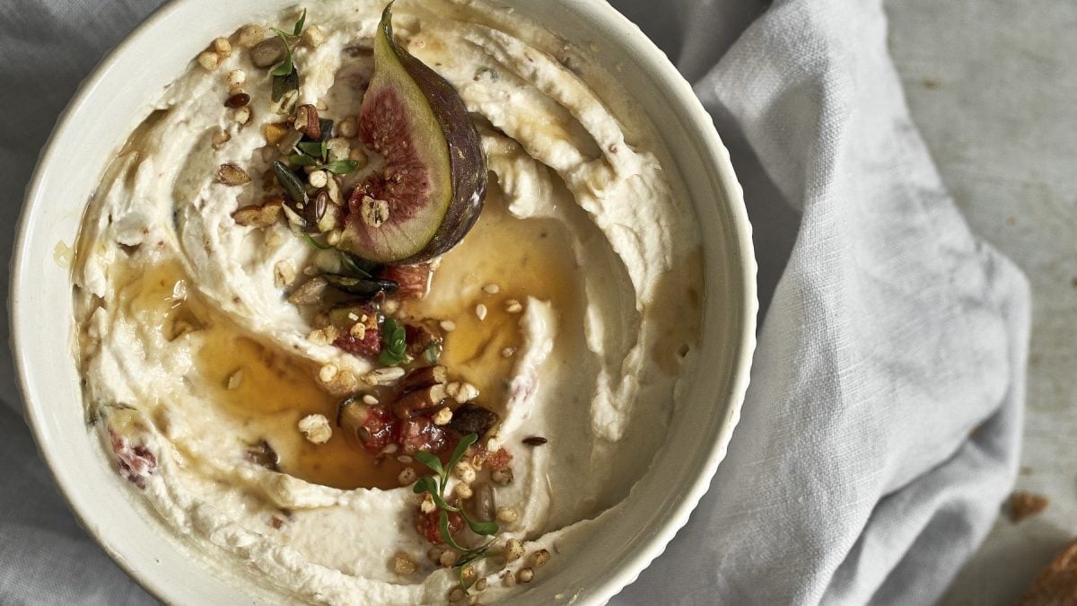 Frischkäsedip mit Feigen und angerösteten Macadamia-Nüssen, übergossen mit Honig und serviert in einer weißen Schale auf hellem Untergrund.