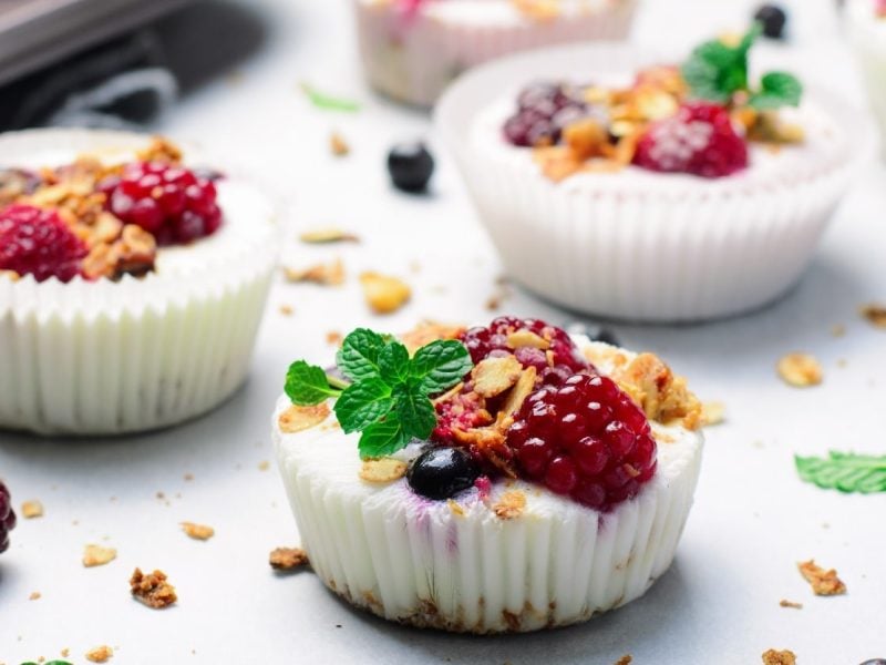 Mehrere Frozen Yoghurt Cups auf einem weißem Tisch. Darauf verteilteinige Haferflocken, Beeren und Minze.