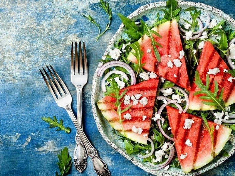 Ein Teller mit grillten Melonen und feta und Rucola steht auf einem blauen Untergrund.