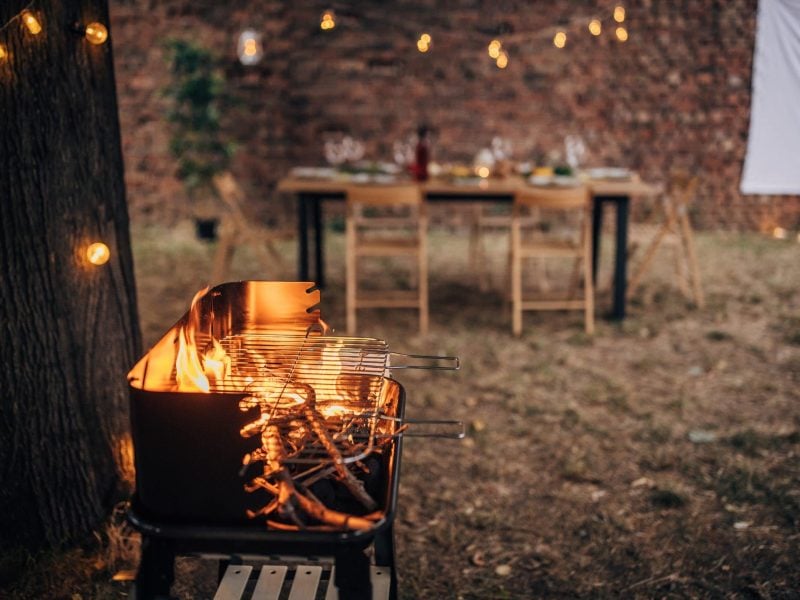 Grillzubehör Basisausstattung 7 Grill-Tools für kleines Geld Grillparty im Hinterhof mit Grill und Flamme. Im Hintergrund Tisch mit Gaben. Frontalansicht.