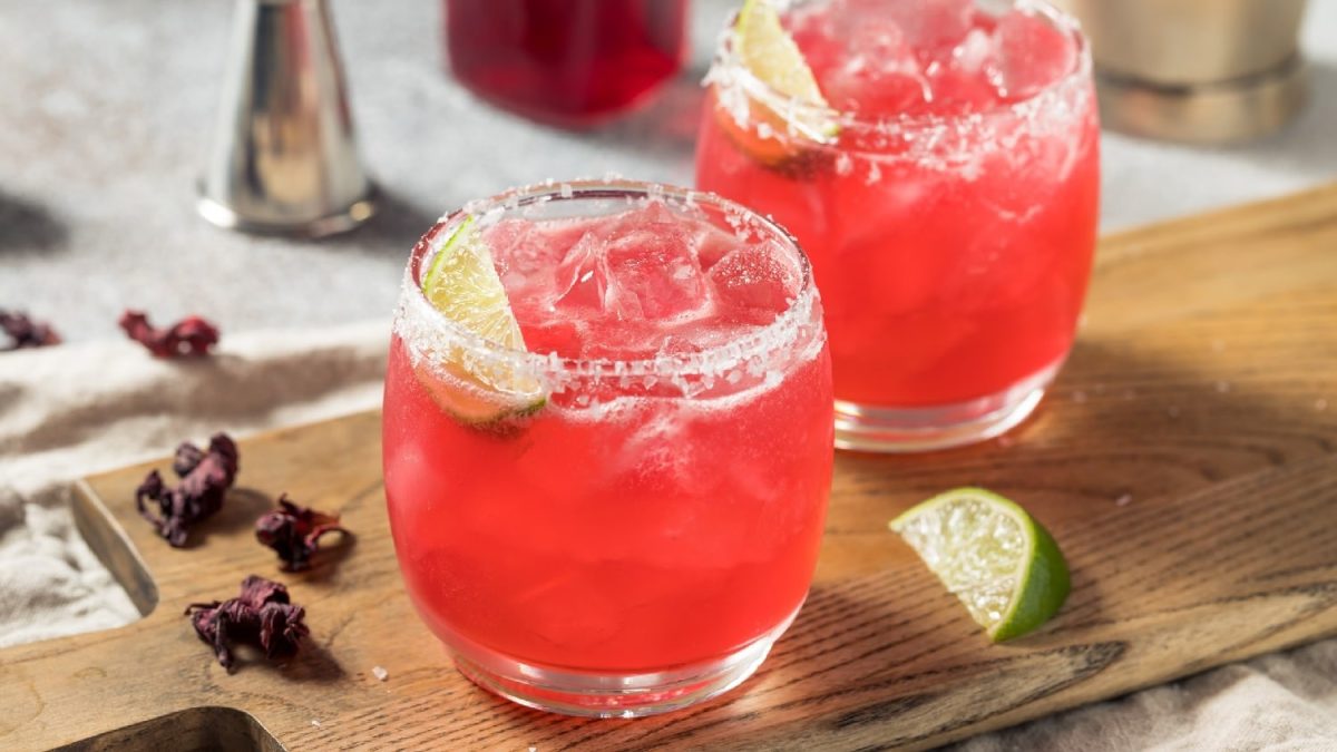 Zwei Gläser mit dem Hibiskus-Mezcal-Margarita auf einem Holzbrett mit Hibiskus-Blüten und Limetten.