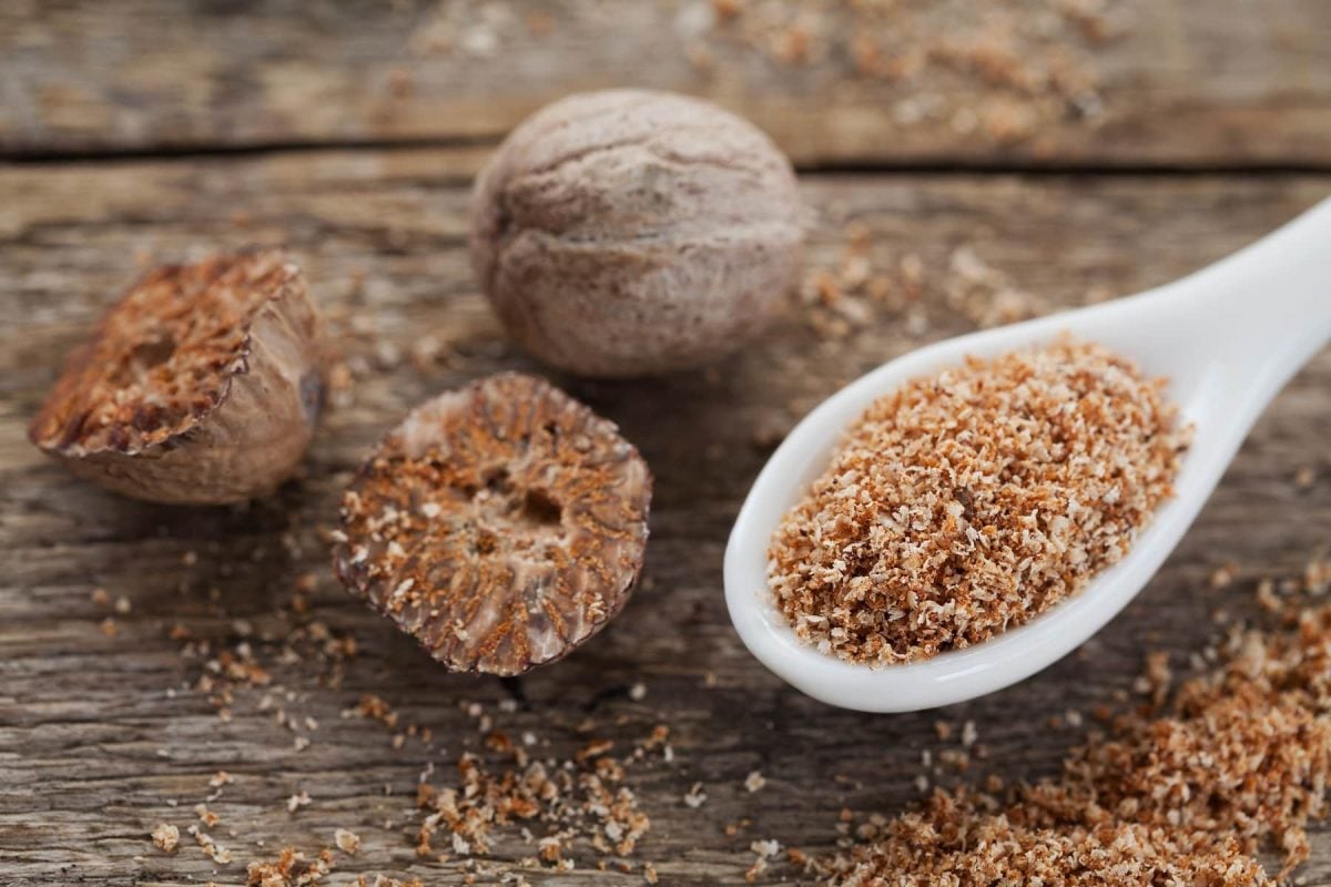 Auf einem Untergrund aus Holz liegen ein ganze Muskatnuss, zwei halbe Muskatnüsse sowie in Löffel mit geriebenem Muskat.