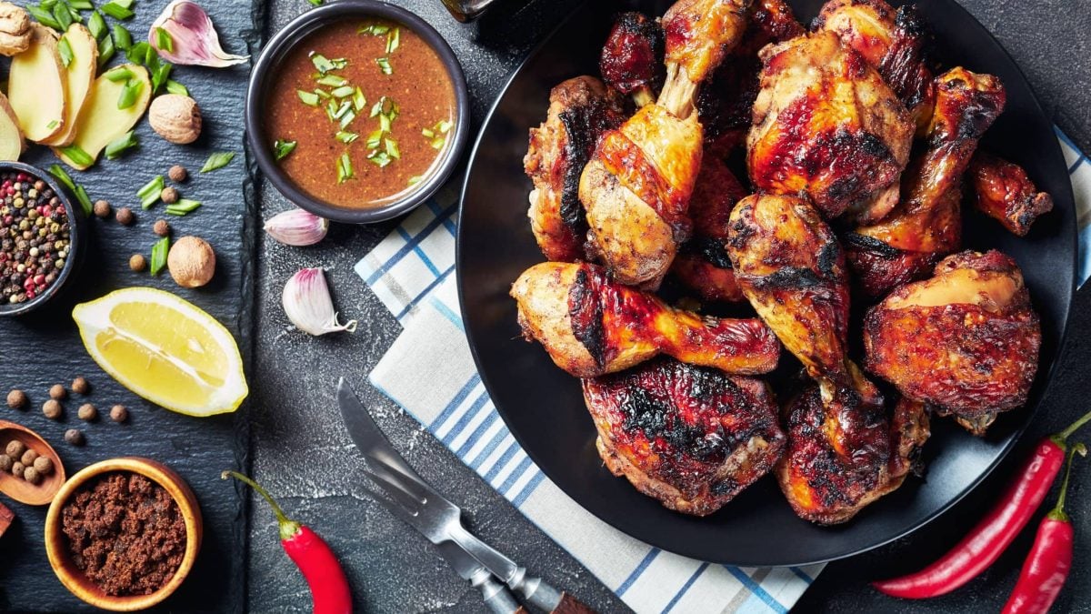 Jerk Chicken auf schwarzem Teller auf Tuch auf grauem Untergrund. Daneben Gewürz, Chilis, Limettenspalten, Knoblauch und Lauchstücken. Draufsicht.und