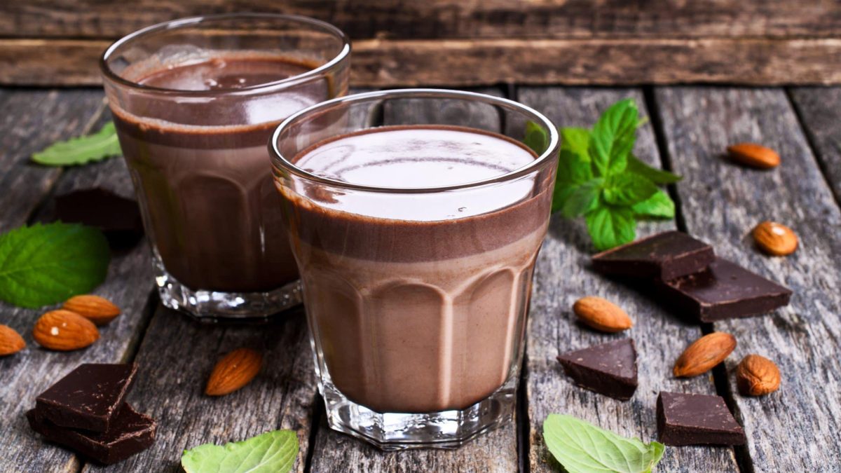 Zweimal Kakao-Shake mit Johannisbeeren und Apfelsaft in Glasern auf grauem Tisch vor Holz. Im Bild Minze, Schokolade und Mandeln. Frontalansicht.