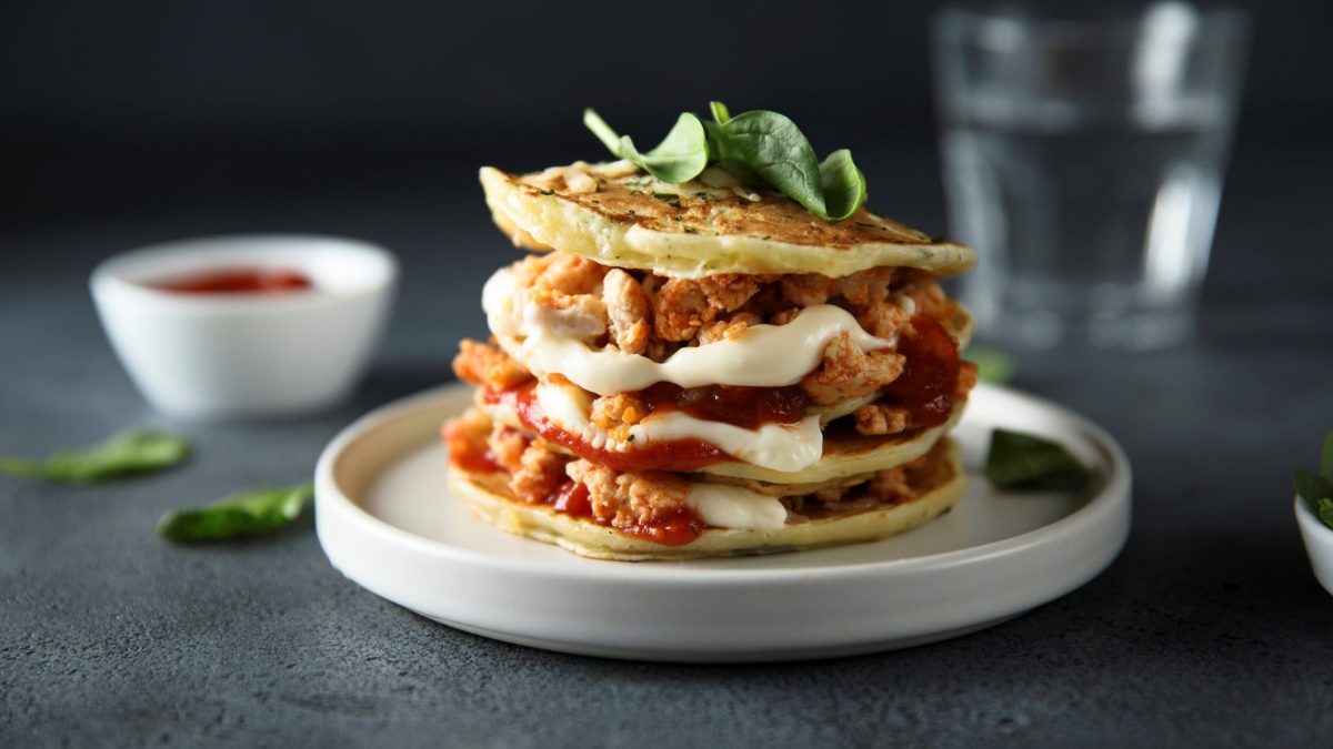 Kräuter-Pancakes mit Hähnchen, Creme und Tomatensauce auf weißem Teller auf grauem Untergrund. Im Hintergrund ein Glas Wasser und Dipschale.