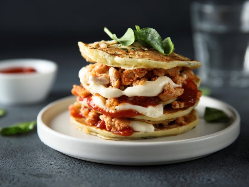 Kräuter-Pancakes mit Hähnchen, Creme und Tomatensauce auf weißem Teller auf grauem Untergrund. Im Hintergrund ein Glas Wasser und Dipschale.