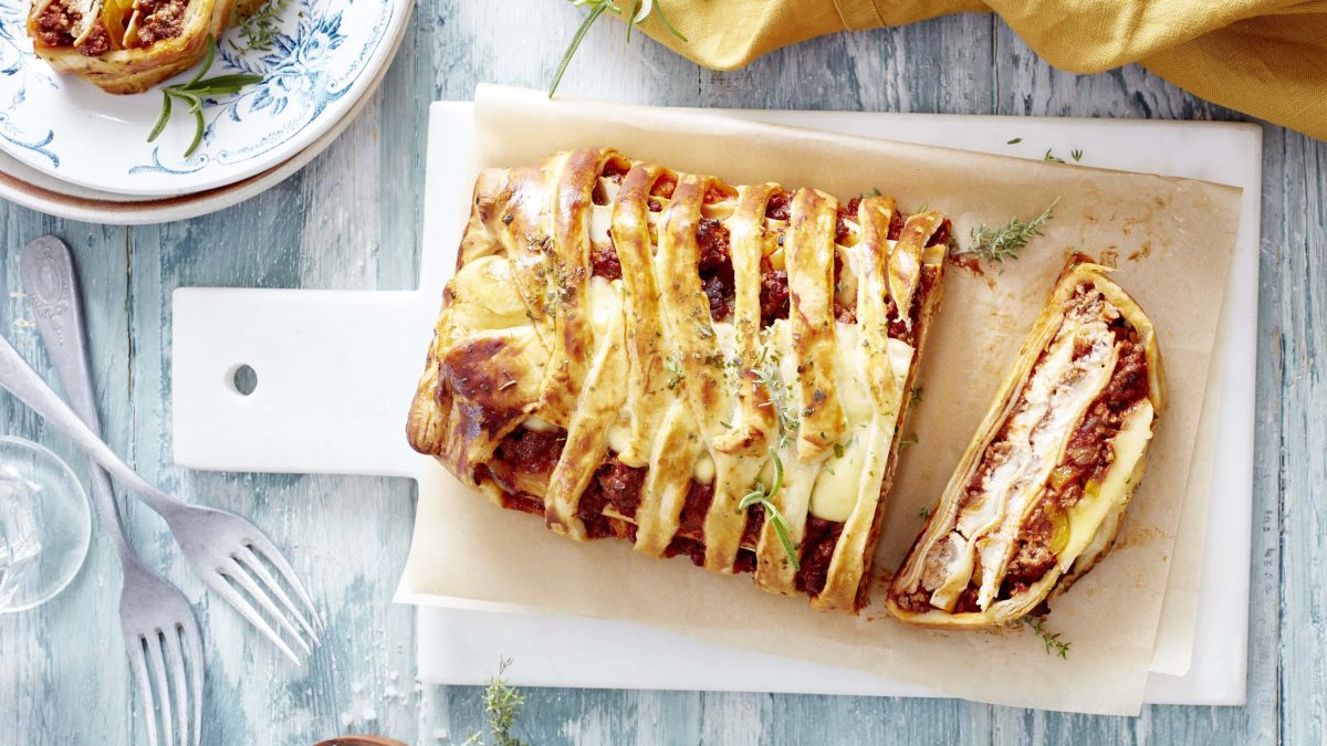 Lasagne-Blätterteigzopf auf Servierbrettchen neben besteck vor hellgrauem Hintergrund