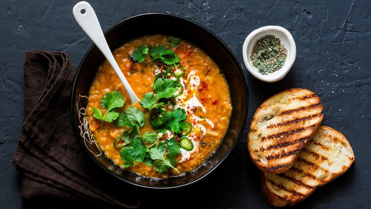 Eine schwarze Schüssel Linsen-Kokos-Suppe mit einem weißen Keramiklöffel darin in der Draufsicht, daneben eine Schale Gewürze, zwei getoastete Brotscheiben und ein braunes Geschirrtuch.