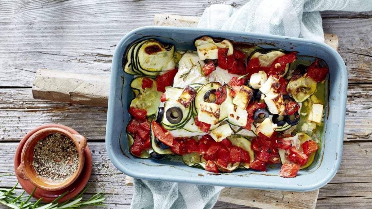 Bunter mediterraner Fischauflauf, bestehend aus leicht geschmorter Zucchini, schwarzen Oliven, Tomaten, gebackenem Feta und saftigem Kabeljaufilet, serviert in einer großen Form auf hölzernem Tisch.