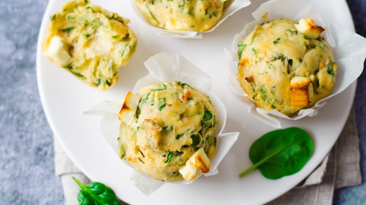Ein weißer Teller auf dem die herzhaften Muffins mit Spinat und Feta liegen. Im Hintergrund ist eine grau marmorierte Wand und etwas Blattspinat zu erkennen.