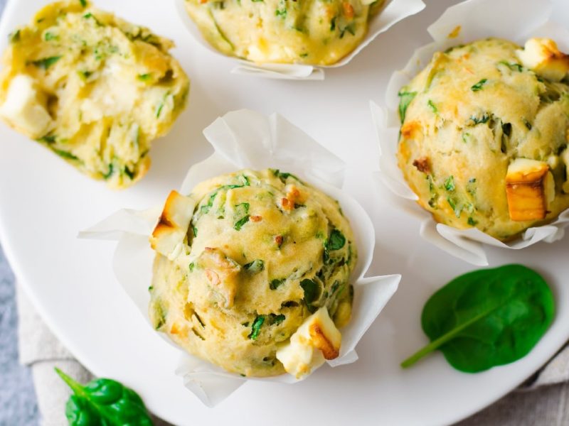 Ein weißer Teller auf dem die herzhaften Muffins mit Spinat und Feta liegen. Im Hintergrund ist eine grau marmorierte Wand und etwas Blattspinat zu erkennen.