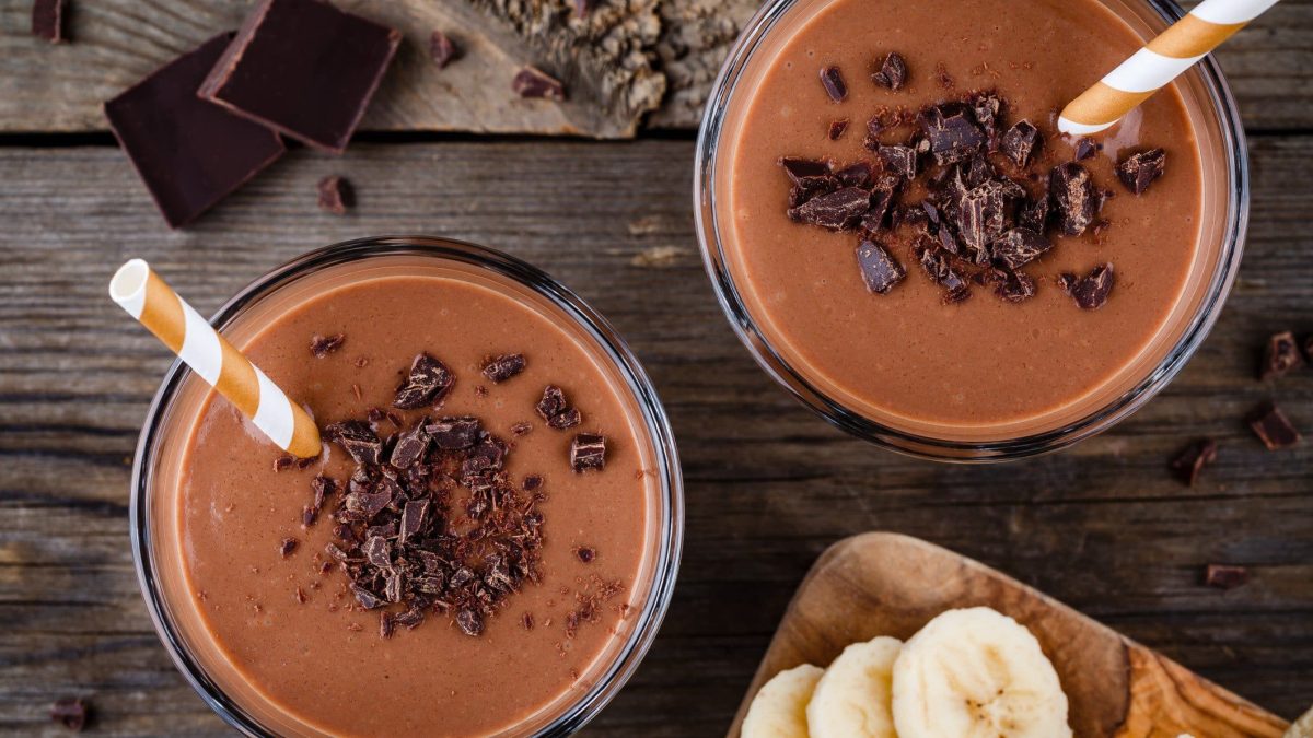 Pistazienmus-Smoothie mit Kakao, Bananen und Beeren in Gläsern mit Strohhalm auf dunklem Holz. Daneben Holzbrett mit Bananenscheiben. Oben Schokolade. Draufsicht.