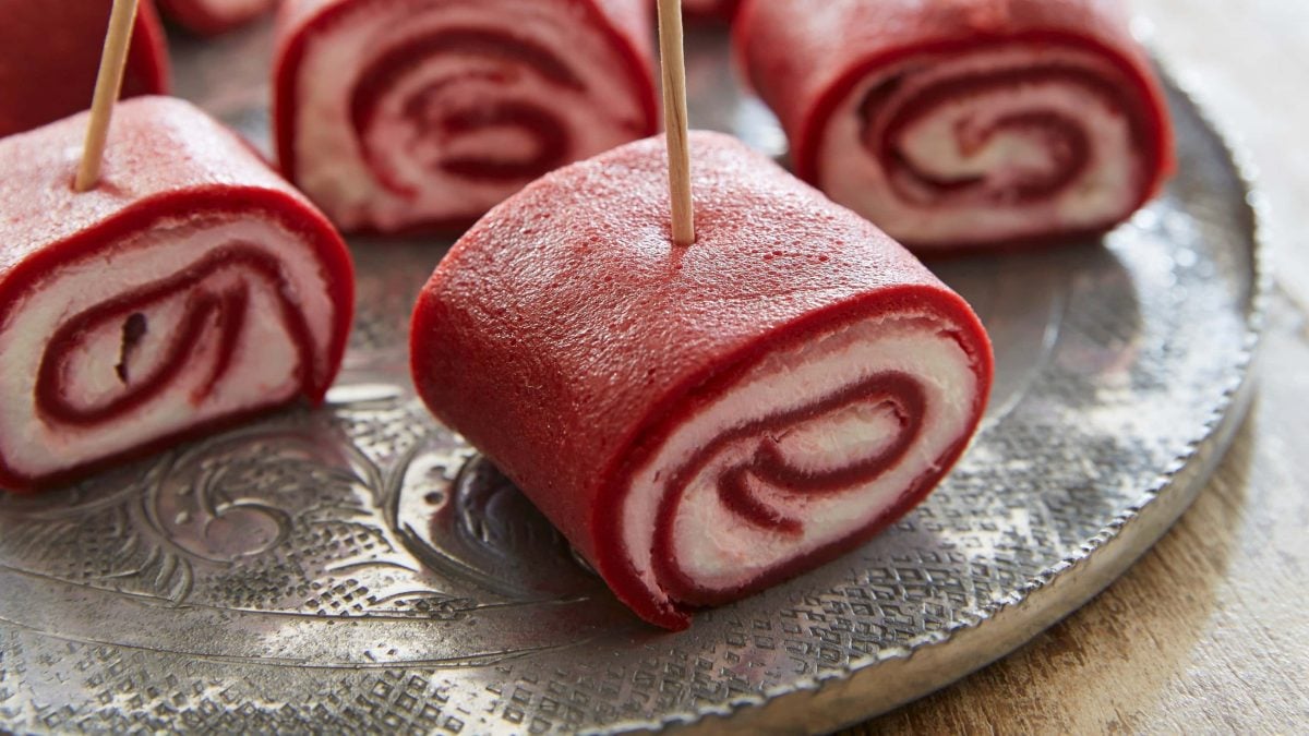Mehrere Rote Bete-Creperöllchen auf kleinen Spießen auf einem dunklen Teller vor Holzhintergrund.