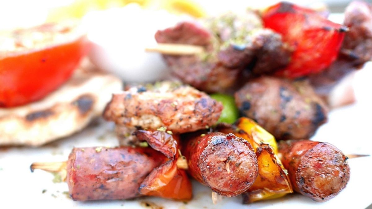 Gegrillte Salsiccia-Spieße mit Gemüse und Chilis auf weißem Untergrund. Daneben Schale mit Dip und Fladenbrot. Frontalansicht.