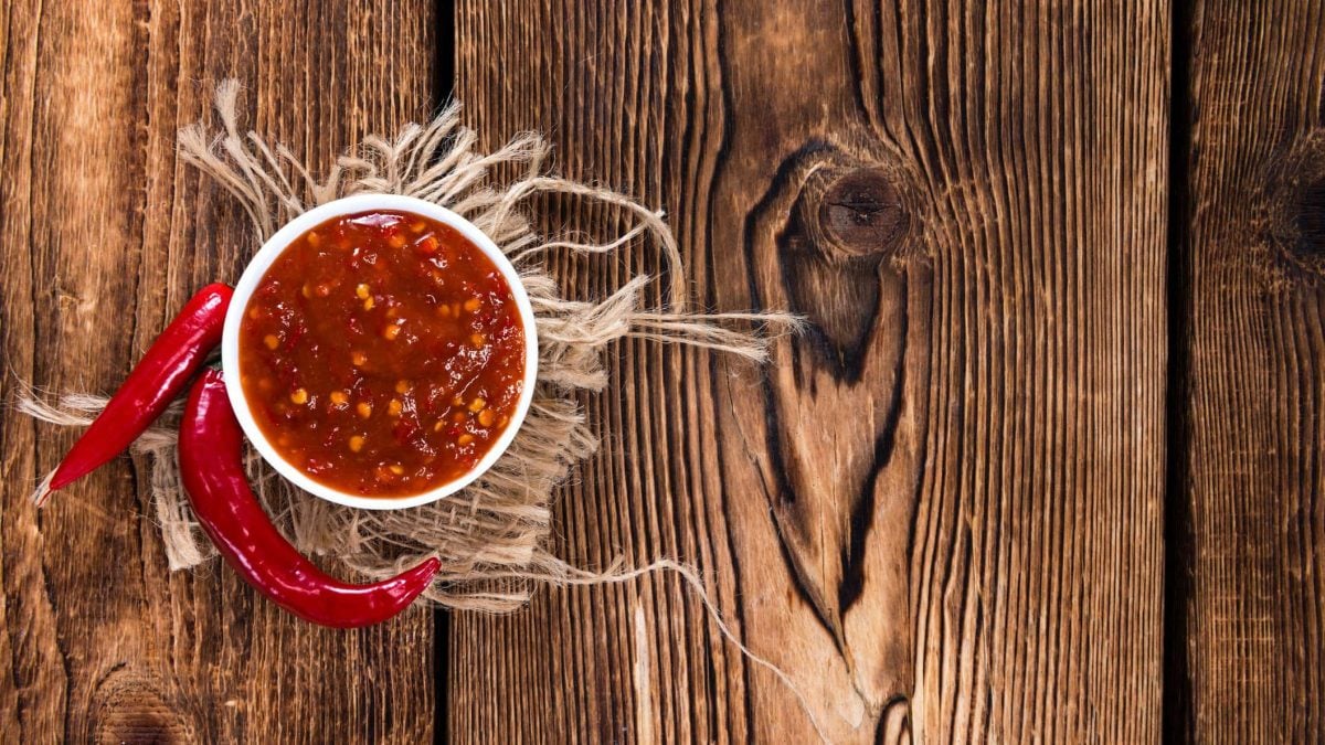 Selbst gemachtes Sambal Oelek in weißer Schale auf beigem Tuch auf Holztisch. Daneben Chilischoten. Aufnahme aus der Vogelperspektive.