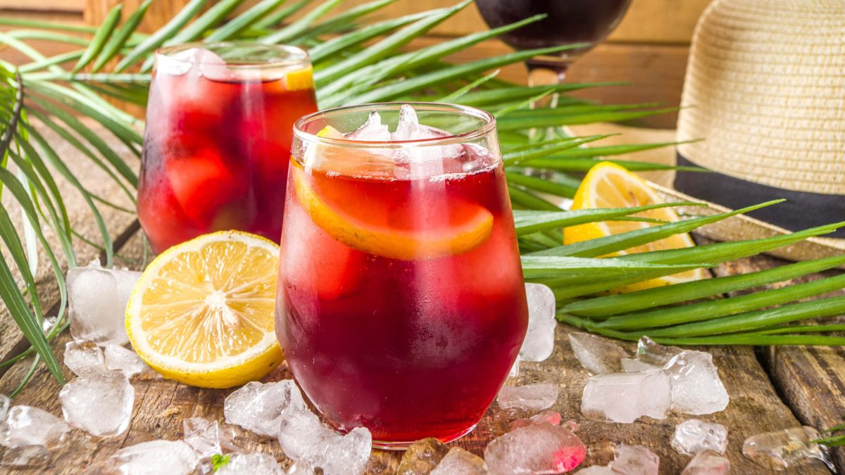 Tinto de Verano in zwei Gläsern auf Holztisch mit Eiswürfeln, Pflanzen, Zitronen und Strohhut. Im Hintergrund Rotweinglas erkennbar. Frontalansicht.