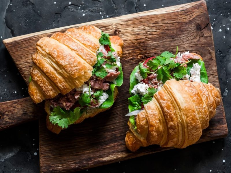 Zwei angeschnittene Croissants auf einem dunklem Holzbrett mit Koriander und Thunfisch.
