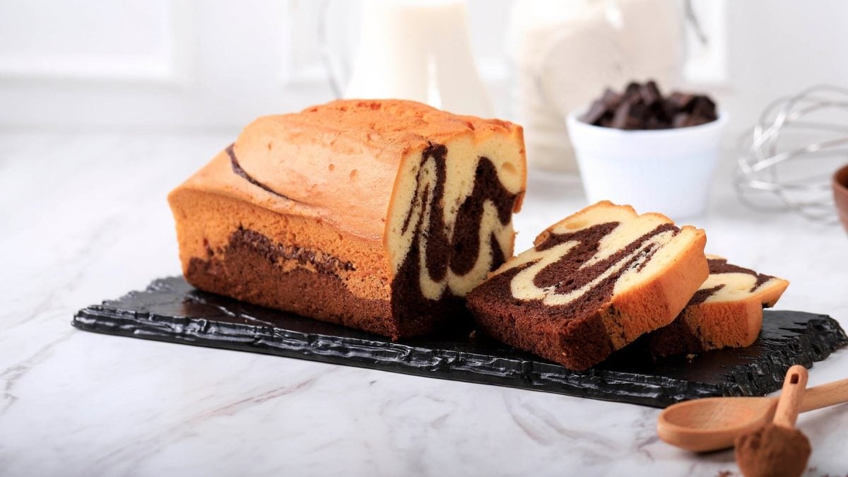 Ein angeschnittener veganer Marmorkuchen auf einer Schieferplatte, im Hintergrund unscharf einige Zutaten und Backutensilien.