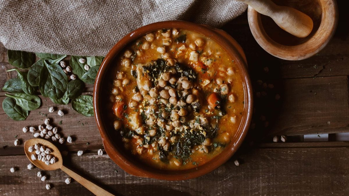 eine Tonschale mit veganem Mangold-Curry mit Kichererbsen in der Draufsicht, daneben Mörser und Stößel sowie ein Holzlöffel und Mangoldblätter.