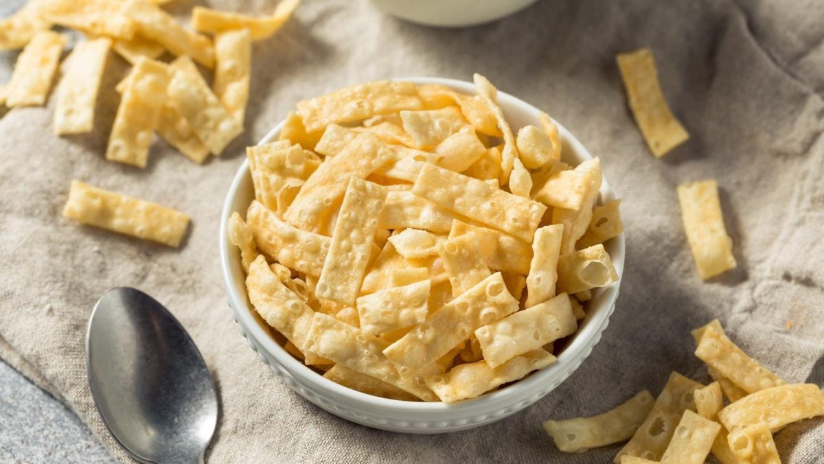 Wantan Chips aus selbst gemachtem Wantan Teig in weißer Schüssel auf Tuch. Im Bild Chips verteilt. Links Metalllöffel. Oben Schale angedeutet. Aufnahme aus der Vogelperspektive.