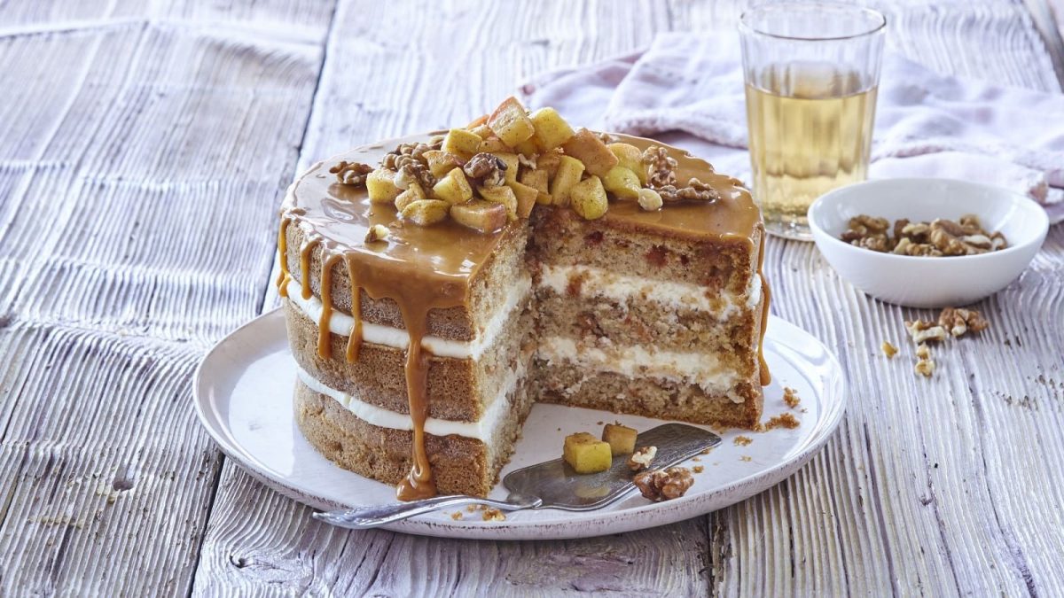 Auf einem Teller steht eine Apfel-Karamell-Schichttorte. Sie ist mit Äpfeln, Zimt und Walnüssen bedeckt.