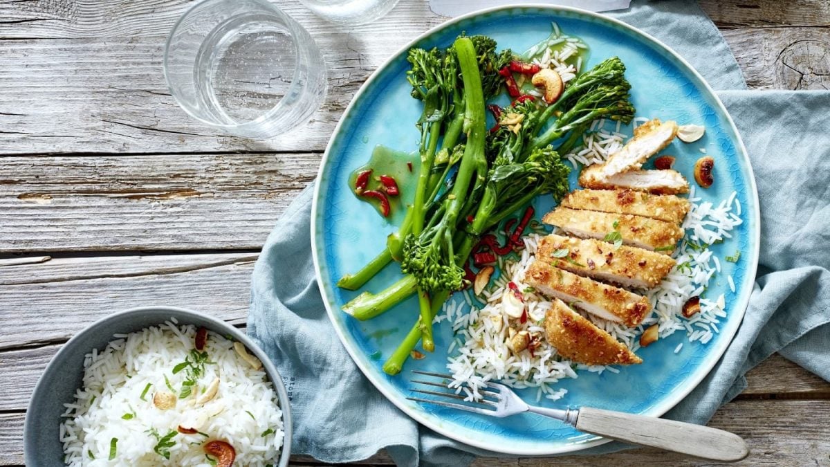 Auf einem blauen Teller liegt ein Hähnchenschnitzel mit Brokkolini. Es wird mit Reis serviert.