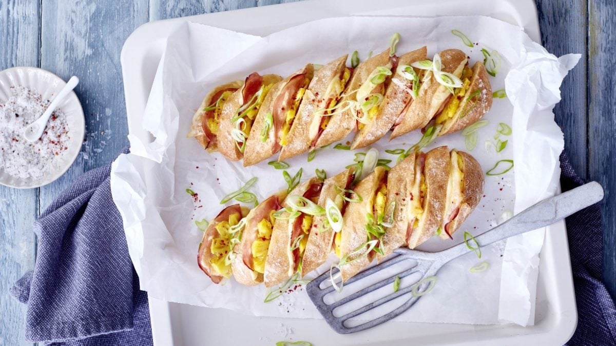 Hasselback-Baguette Hawaii, serviert in einer weißen Auflaufform mit Backpapier, garniert mit Frühlingszwiebeln und frischen Pfeffer- und Salzkörnern auf grauem Untergrund.
