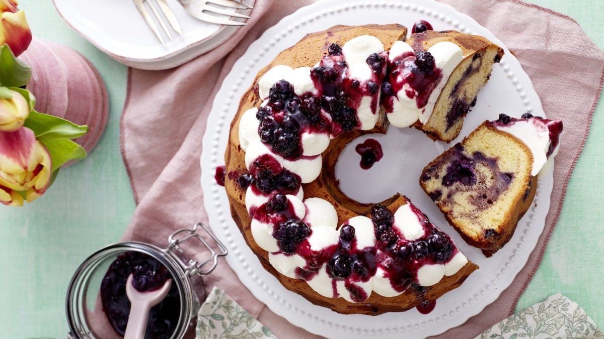 Mit Sahne und Heidelbeerkonfitüre garnierter Heidelbeer-Marmorkuchen, serviert auf einem weißen Teller auf rosanem Tischtuch mit zusätzlicher Konfitüre und einem Strauß Tulpen.