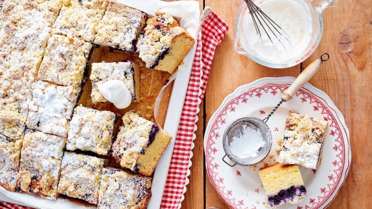 Heidelbeerkuchen mit Orangenstreuseln, als Blechkuchen in kleine Stücke angeschnitten auf einer Form serviert, mit Puderzucker bestreut und mit geschlagener Sahne gereicht.