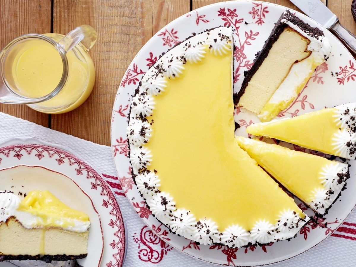 Kakaokeks-Käsekuchen, bestehend aus einer Eierlikörkäsekuchencreme, einem Kakaokeksboden und einer Garnitur aus kleinen Sahnetupfern und Eierlikörglasur, serviert auf einem rot-weißen Teller mit Blumenmuster auf einem Holztisch.