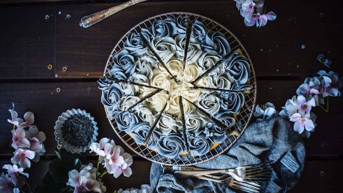 Auf einem Kuchenteller steht ein Kuchen mit schwarzem Sesam und Vanille. Er ist mit Creme dekoriert, die in Rosen angeordnet ist und einen Farbverlauf ergibt.