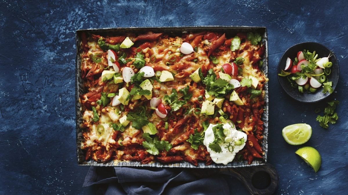 Nudel-Hühnchen-Auflauf, überbacken mit Käse und mit selbst gemachter Salsa garniert, in der Form auf einem schiefernen Tisch serviert mit frischen Limetten und Sour Cream.